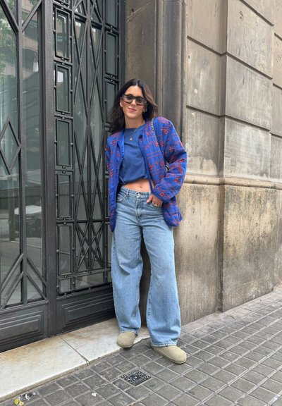 Chaqueta azul con un patrón de diseños rojos, parte superior azul y jeans de pierna ancha de mezclilla clara. Zapatos beige redondeados. De pie junto a una puerta de vidrio con patrón.