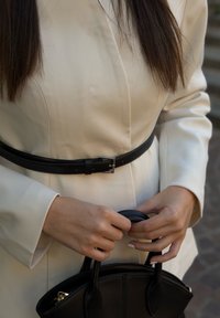 Sacou crem cu mâneci lungi și o curea neagră, ținând o geantă neagră structurată. Material neted și texturi contrastante.