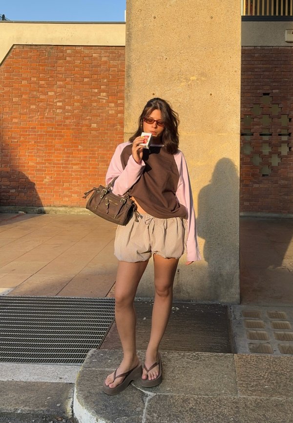 Jeune femme portant des lunettes de soleil, un t-shirt à manches longues marron et rose, un short et des sandales, sirote une boisson debout près d'un pilier en béton à l'extérieur.