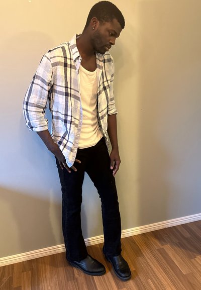 Camisa de botones de cuadros en blanco, azul y negro sobre una camiseta blanca, combinada con jeans negros y botas negras, de pie sobre un suelo de madera.