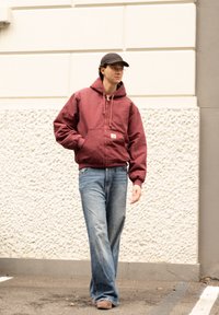 Καφέ-κόκκινη φούτερ μπλούζα με κουκούλα και μπροστινές τσέπες, συνδυασμένη με ανοιχτό γαλάζιες τζιν. Το μοντέλο φοράει μαύρο καπέλο και περπα�τάει μπροστά από έναν υφασματοποιημένο τοίχο.