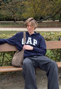 Marineblå hættejakke med "CAP" bogstaver, grå oversized bukser og beige skuldertaske, siddende på en træbænk i en park.