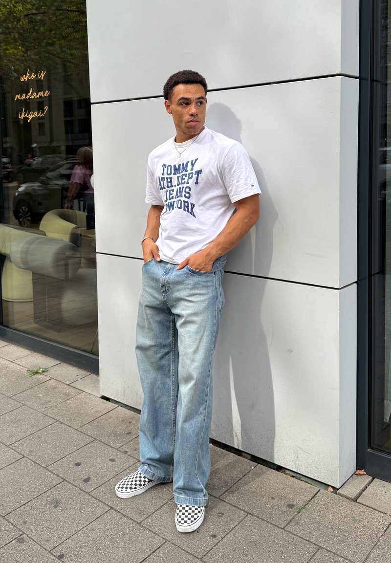 Jeune homme en t-shirt blanc à motif graphique, jean bleu clair et chaussures slip-on à damier, appuyé contre un mur blanc d’un bâtiment moderne.