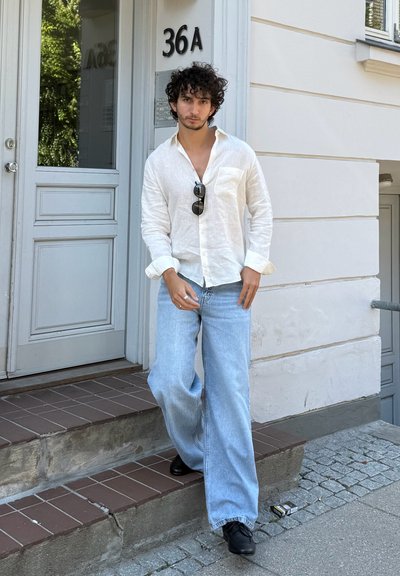 Camisa blanca de lino con botones, jeans de pierna ancha azul claro y zapatos negros. De pie en unos escalones cerca de una puerta gris marcada con "36A".