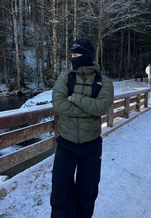 Persona con chaqueta acolchada verde y cubrebocas negro, de pie con los brazos cruzados en un puente de madera nevado junto a una orilla de río boscosa y cubierta de nieve.