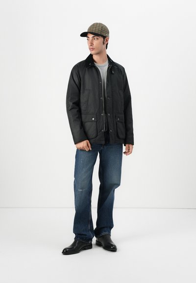 Chaqueta negra acolchada de botones con dos bolsillos frontales, llevada sobre una camiseta gris. Combinada con jeans azules y botas de cuero negras, rematada con una gorra de cuadros.