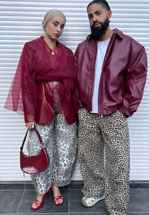 Chaqueta de cuero roja sobre una camisa blanca; top transparente granate; pantalones gris con estampado de leopardo; sandalias rojas; bolso burdeos; fondo con persianas blancas.