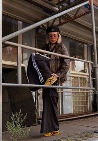 Barna bőrkabát, fekete nadrág fehér csíkokkal, sárga sportcipő, fekete beret, és napszemüveg. Áll egy állvány háttér előtt.