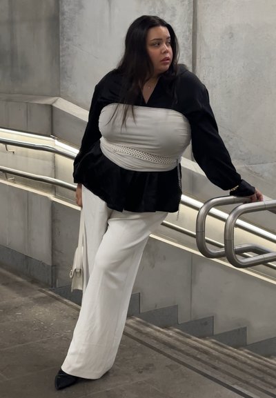 Mujer de cabello largo y oscuro apoyada en una barandilla de metal junto a unas escaleras de concreto, vistiendo blusa negra, cinturón corsé blanco, pantalones blancos y sosteniendo un bolso blanco.