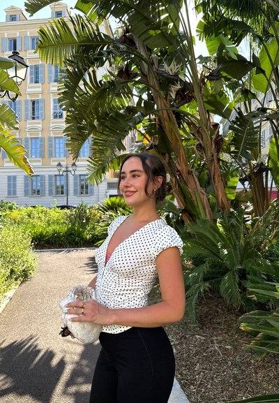 Blusa blanca de manga corta con lunares negros, diseño ajustado, combinada con pantalones negros. En el fondo hay follaje verde y un edificio.