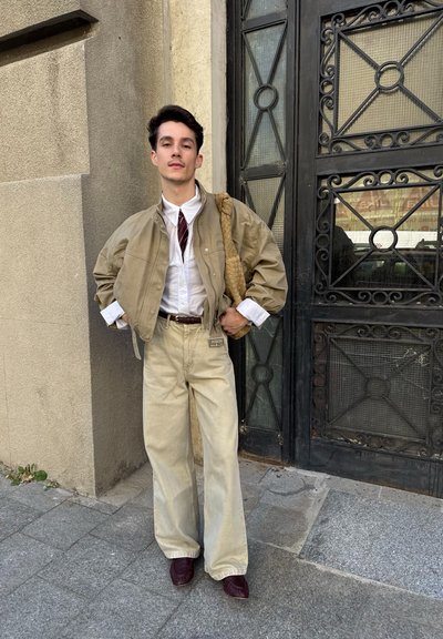 Joven con cabello oscuro lleva chaqueta beige, camisa blanca, corbata roja, pantalones beige anchos y zapatos oscuros, de pie junto a una puerta decorativa de metal negro.