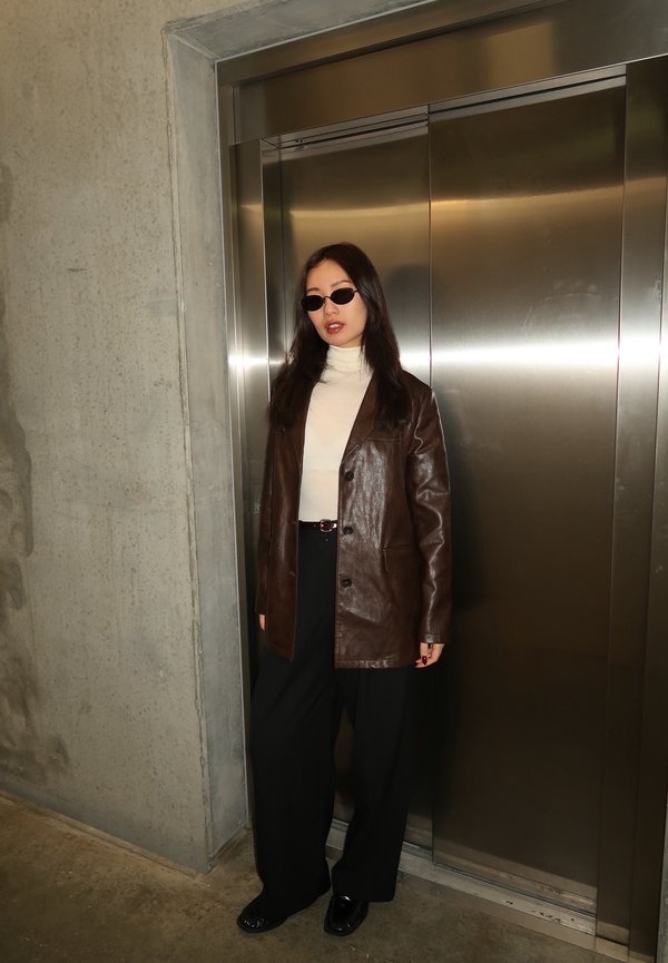 Woman wearing sunglasses, a brown leather jacket, a white roll-neck jumper, and black wide-leg trousers stands in front of closed metallic lift doors.