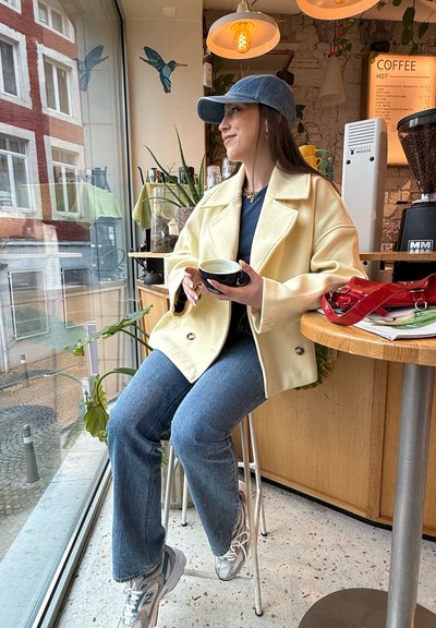 Abrigo oversized color crema con grandes solapas, gorra de denim azul, camiseta azul marino y jeans azul claro; taza negra en la mano, zapatillas plateadas.