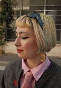 Różowa koszula z krótkim rękawem, wzorzysty krawat, szary sweter, złote kolczyki w kształcie serca, warstwowe pastele na włosach, mocny eyeliner i kolczyki w nosie.