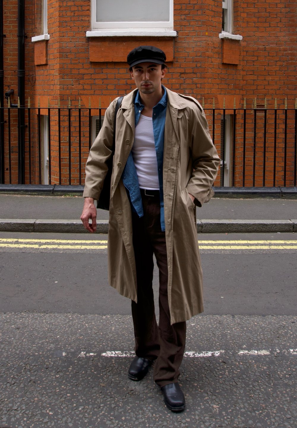 Beige trench coat over en blå skjorte og en hvit tank topp. Brune bukser og svarte sko. Svart caps. Urban gatebakgrunn med murvegg.