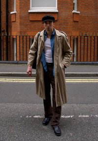 Beige trench coat over en blå skjorte og en hvit tank topp. Brune bukser og svarte sko. Svart caps. Urban gatebakgrunn med murvegg.