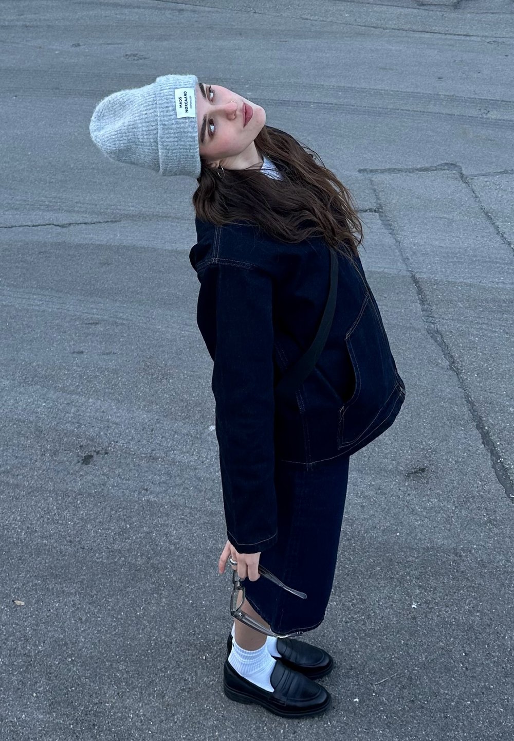 Μοντέλο φοράει ένα σκούρο τζην μπουφάν, αντίστοιχη φούστα, γκρι σκουφάκι και μαύρες λαστιχένιες γόβες. Κρατάει γυαλιά και στέκει σε μια γκρίζα πλακόστρωτη επιφάνεια.