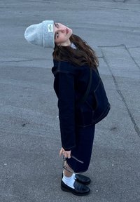 Μοντέλο φοράει ένα σκούρο τζην μπουφάν, αντίστοιχη φούστα, γκρι σκουφάκι και μαύρες λαστιχένιες γόβες. Κρατάει γυαλιά και στέκει σε μια γκρίζα πλακόστρωτη επιφάνεια.