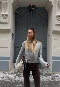 Jeune femme aux longs cheveux blonds portant un pull gris et un pantalon noir, tenant un sac beige, debout devant une porte ornée gris foncé numérotée 29.