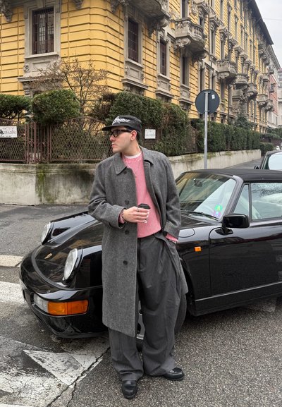 Hombre con abrigo gris, suéter rosa y gorra sostiene una taza de café junto a un convertible negro vintage en una calle de la ciudad con un edificio amarillo de fondo.