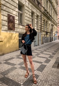 Melnā džinsa jaka, tirkīza kreklis, melni īsie šorti un brūnas siksnu sandales. Tur rokās saulesbrilles, ar rakstainu soma. Bruģēta iela fons.