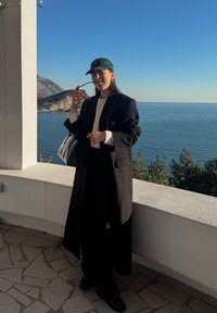 Femme en manteau noir et casquette tenant des lunettes de soleil sur une terrasse donnant sur une mer calme et une côte montagneuse baignée de lumière solaire.