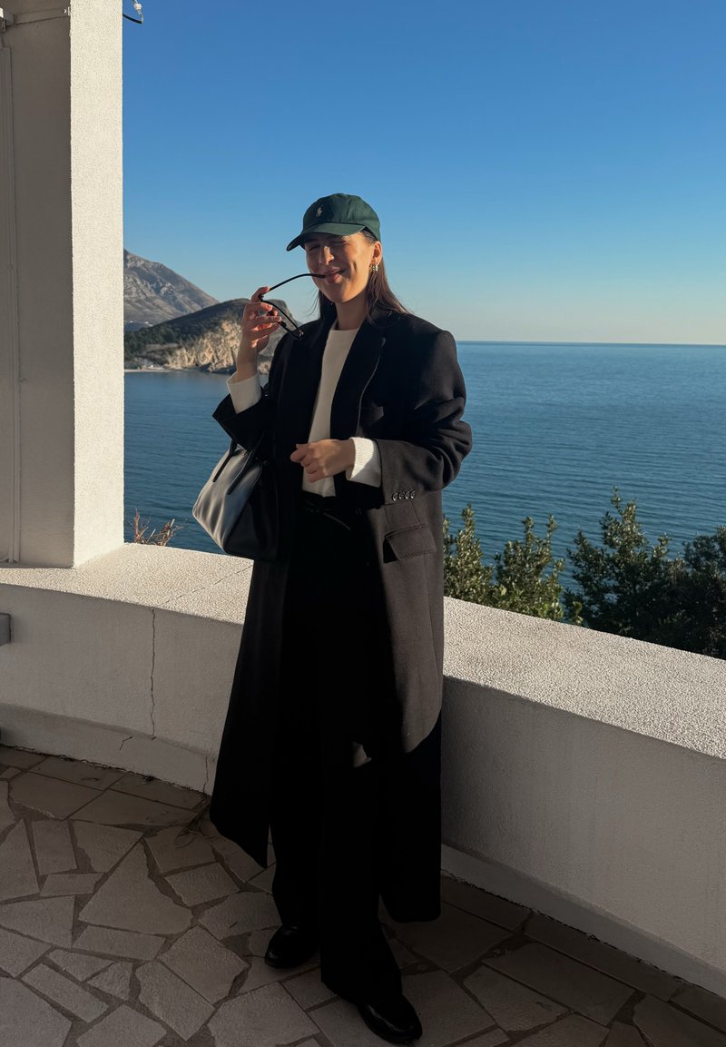 Femme en manteau noir et casquette tenant des lunettes de soleil sur une terrasse donnant sur une mer calme et une côte montagneuse baignée de lumière solaire.