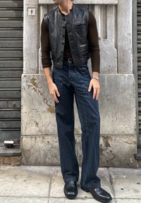 Moški nosi črn usnjen jopič čez dolgo-sleeve rjav srajco, široke hlače z visokim pasom in črne čevlje s kvadratnim prstom, stoji na kamnitih ploščicah.