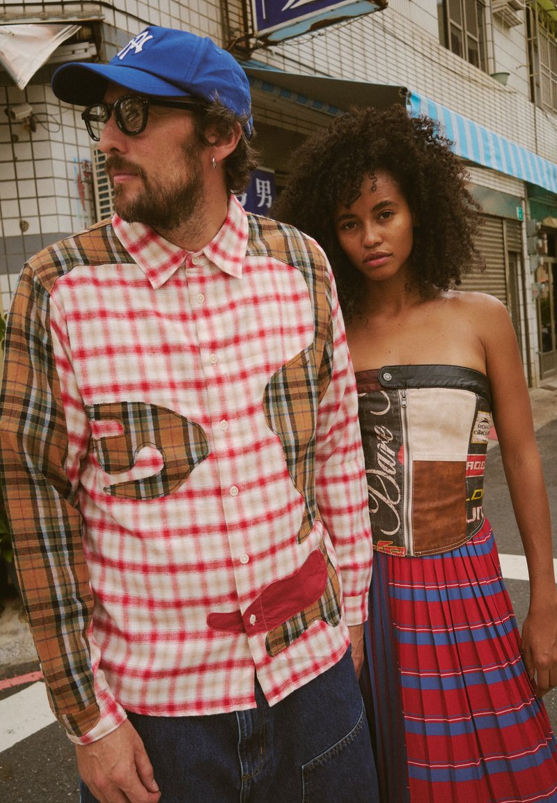 Camisa xadrez com padrões vermelhos e castanhos, abotoada, combinada com uma saia listrada colorida e um corpete de retalhos, ao ar livre.