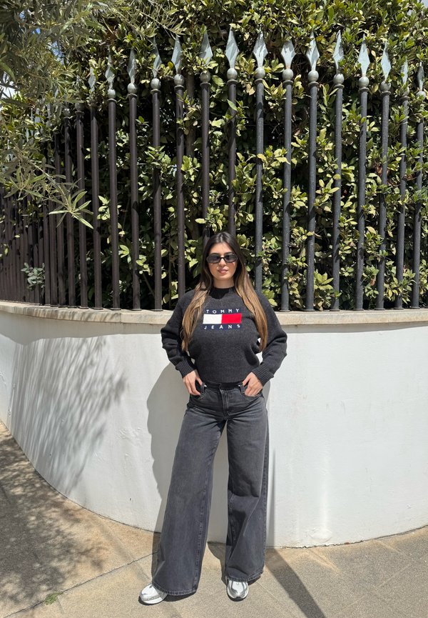 Γυναίκα με γυαλιά ηλίου, μαύρο πουλόβερ Tommy Jeans, φαρδιά σκούρα τζιν και λευκά αθλητικά παπούτσια στέκεται με τα χέρια στις τσέπες δίπλα σε λευκό τοίχο και μαύρο φράχτη με πράσινα φυτά.