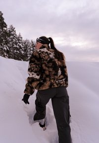 Valse bontjas met zwart-bruine abstracte patronen, gecombineerd met donkere sneeuwbroek. Handschoenen aan; besneeuwd landschap op de achtergrond.