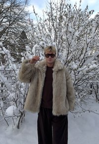 Manteau en fausse fourrure beige clair avec une texture pelucheuse et de larges revers, porté sur un haut foncé et un pantalon noir ample, sur fond de paysage enneigé.