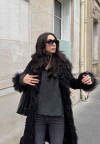 Žena s dlhými tmavými vlasmi, ktorá má na sebe čierne slnečné okuliare, čierny kabát lemovaný kožušinou, tmavý sveter a tmavé džínsy, stojí na mestskom chodníku.