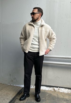 Chaqueta de forro polar con cremallera en crema, superpuesta sobre un cuello de tortuga blanco, combinada con pantalones negros y zapatos negros; diseño minimalista.