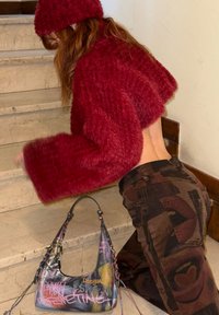 Pull court bordeaux en tissu duveteux avec manches larges, associé à un pantalon marron à motifs. Un sac à bandoulière multicolore de style graffiti est à proximité.