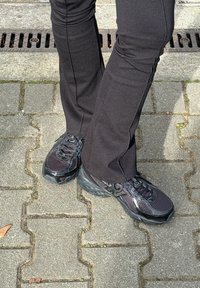 Jambes en pantalon noir et chaussures de sport noires, debout sur un pavé gris à emboîtement près d'une grille d'évacuation métallique.