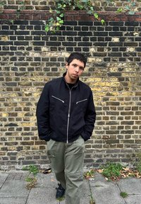 Mladic s kratkom tamnom kosom u crnoj jakni i zelenim cargo hlačama stoji naslonjen na stare opečene zidove, obložene puzavicama i raspršenim lišćem.