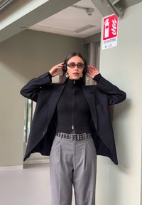 Fekete oversize blézer, bordázott fekete felső, szürke pliszírozott nadrág, tüskés öv, napszemüveg és fejhallgató. Minimalista környezet biztonsági táblával.