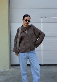 Camisa marrón de gran tamaño con cuello atado, combinada con jeans de color azul claro y una bolsa beige. La camisa tiene una textura suave.