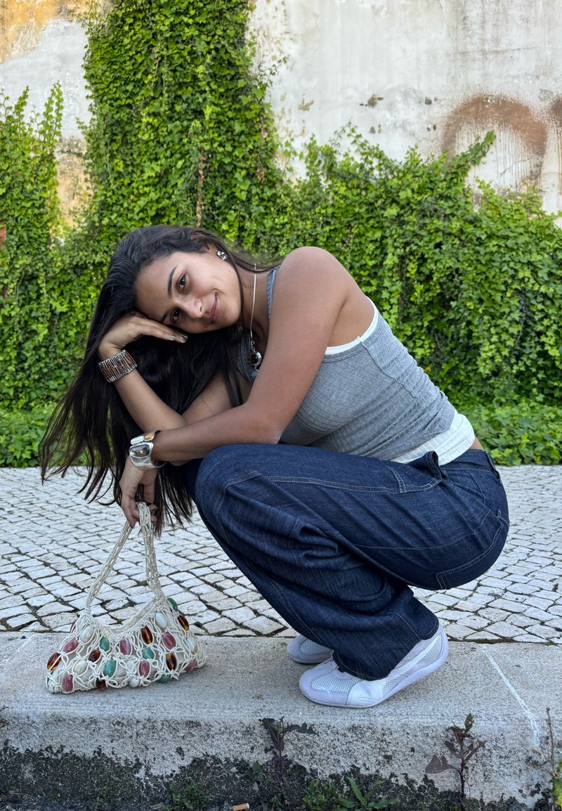 Jeune femme accroupie sur un trottoir, vêtue d'un débardeur gris, jean foncé, baskets blanches, tenant un sac en filet avec des perles colorées, fond de lierre vert.