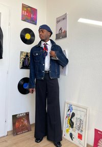 Denim jakna, bela srajca, črtasta kravata in široke črne hlače. Stoji pred vinilnimi ploščami in okvirjenimi umetninami na steni.