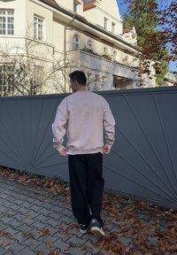 Lichtroze sweatshirt met een losse pasvorm, gecombineerd met zwarte wijde broek. Achteraanzicht tegen een grijze geometrische poort, met gevallen bladeren in de buurt.