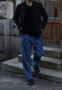 Blazer escuro sobre um suéter preto, combinado com jeans denim azuis folgados e sapatos pretos. A figura está a descer escadas de pedra.