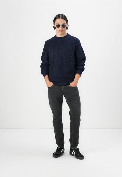 Jersey de punto azul marino con un patrón de trenzas texturizado, jeans grises y zapatillas negras con acentos blancos. El modelo lleva gafas de sol redondas.
