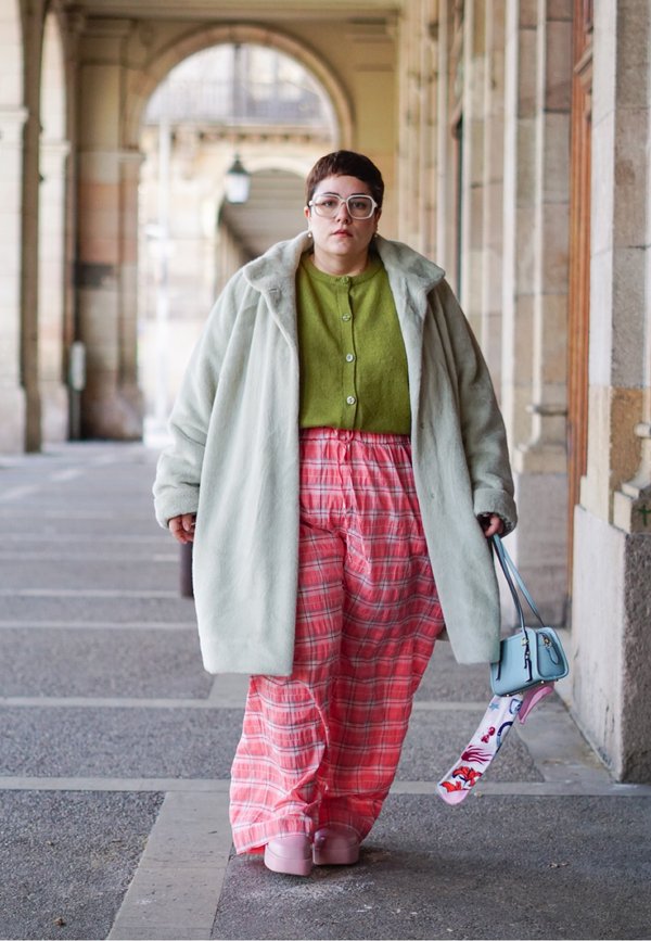 Persona con cabello corto que lleva un abrigo gris claro de gran tamaño, suéter verde con botones, pantalones de cuadros rosas, zapatos rosas y lleva un bolso azul con una bufanda estampada.