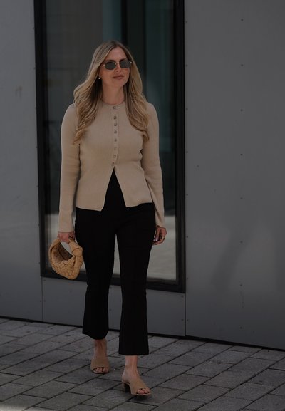 Mujer con cabello largo y rubio, que lleva un suéter beige abotonado, pantalones cortos negros, gafas de sol, sandalias beige con tacón y lleva un bolso tejido caminando al aire libre.