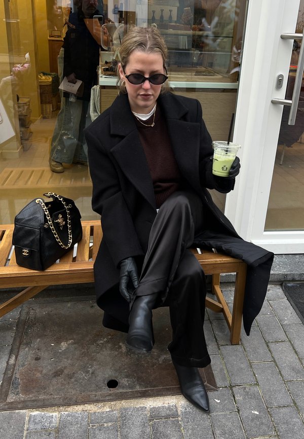Vrouw in een zwarte jas, zonnebril en handschoenen zit op een houten bankje buiten, terwijl ze een groene drank met een rietje vasthoudt naast een zwarte handtas.