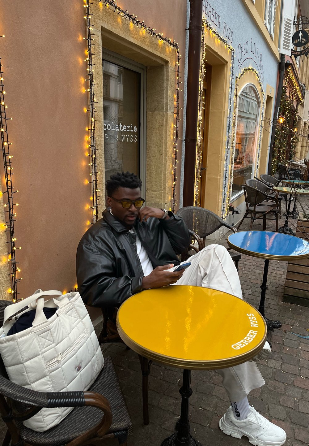 Giacca di pelle nera, tavolo giallo con logo, borsa a tracolla bianca e una persona seduta con uno smartphone in un caffè all'aperto con pavimentazione in ciottoli.