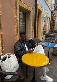 Giacca di pelle nera, tavolo giallo con logo, borsa a tracolla bianca e una persona seduta con uno smartphone in un caffè all'aperto con pavimentazione in ciottoli.