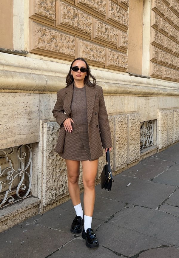 Mujer con blazer y falda marrón a cuadros, top marrón, calcetines blancos, zapatos negros y gafas de sol, de pie junto a una pared de piedra texturizada con un bolso negro.